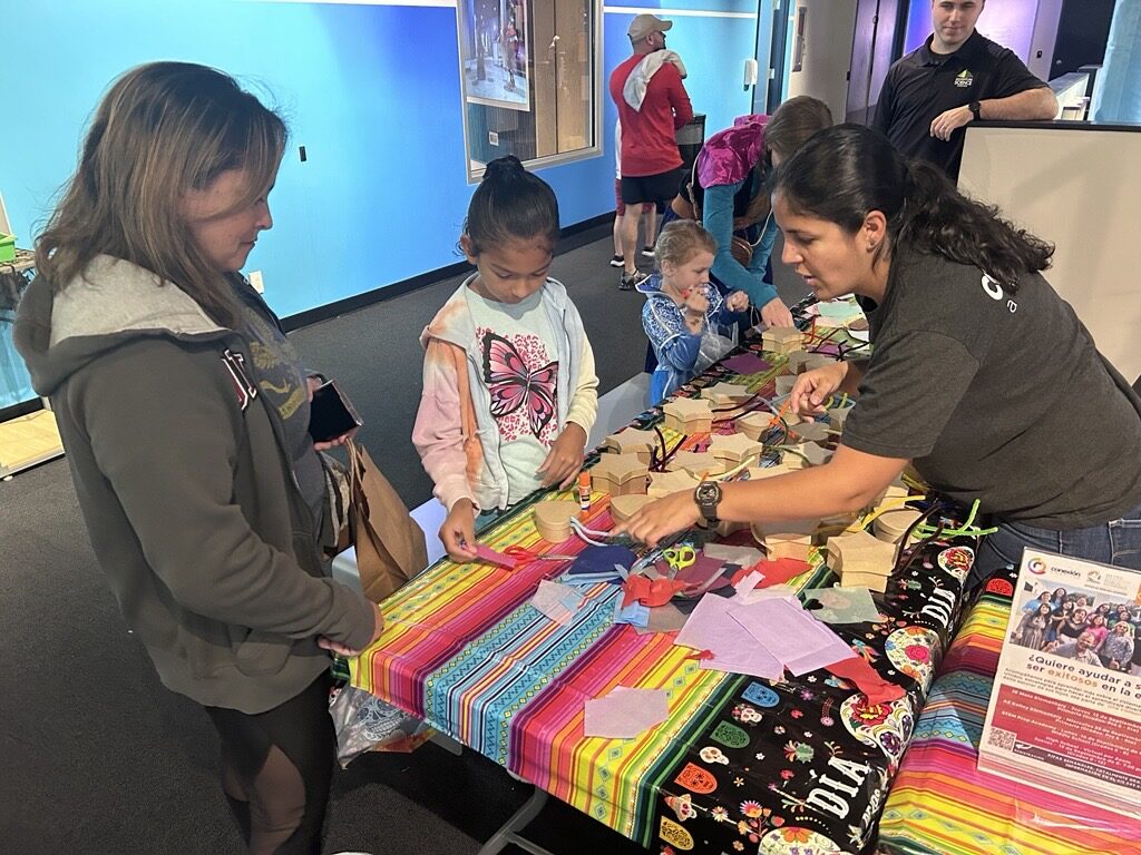 Families participate in a Día de los Muertos craft activity at Adventure Science Center, creating colorful decorations at a table covered with festive fabric in partnership with Conexión Américas.